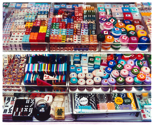 Photograph by Richard Heeps. Three compartmentalised shop shelves, each compartment has coloured plastic dice, counters, circular containers, cards, poker chips, and other plastic type games items.