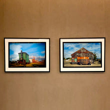 Load image into Gallery viewer, Two framed landscape photographs by Richard Heeps hanging side-by-side on a beige wall. The left frame shows a green steam train on a track under a blue sky, and the right frame shows an old weathered barn with a rusted orange truck under a blue sky.