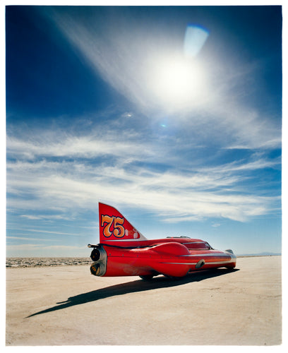 Photograph by Richard Heeps. A red drag car with a 75 written on its fin sits on a salt plain the front facing away towards the right. A blue cloudy sky is overhead.