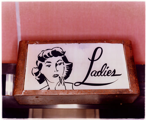 Photograph by Richard Heeps. A kitsch Ladies' toilet sign. The sign has the word Ladies alongside an outline of 1950s woman. The sign sits in a wooden frame against a pink tiled wall.