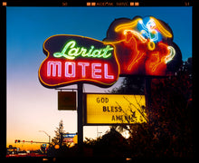 Load image into Gallery viewer, Photograph by Richard Heeps. The photograph is taken in the evening, a neon sign belonging to the Lariat Motel, with a cowboy on horseback with a lasso on the right of the words, half way down the sign post is the yellow sign God Bless America.