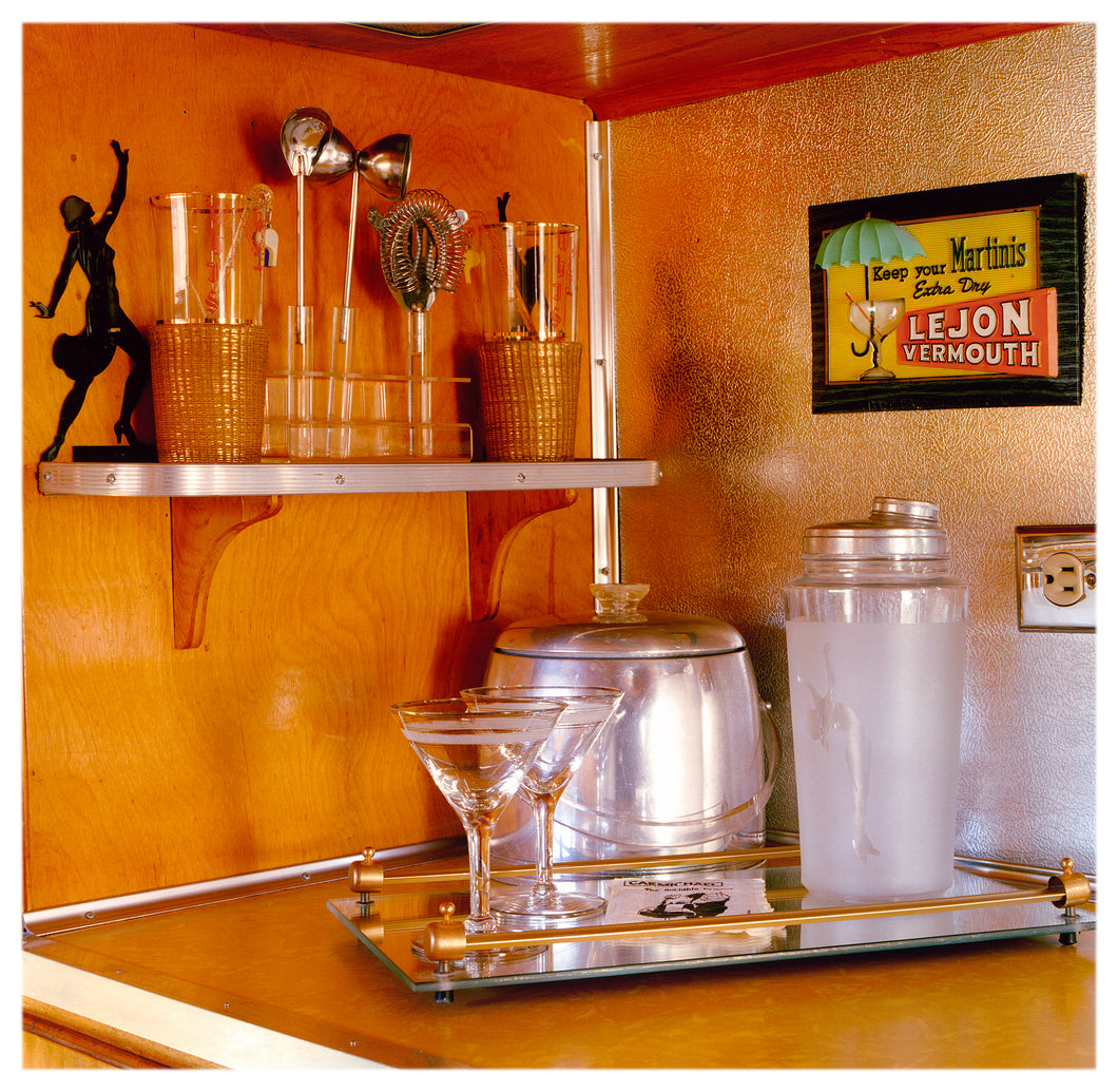 Photograph by Richard Heeps. A drinks corner with martini glasses, cocktail shaker and ice bucket. There is a shelf with cocktail making equipment and it is set against retro orange walls.