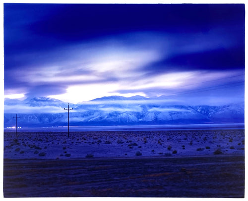 Photograph by Richard Heeps. A blue light hits vast land, mountains and a big sky.