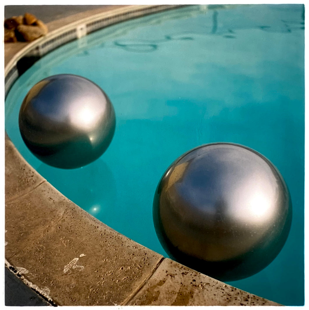 Photograph by Richard Heeps. The corner of a circular swimming pool with two metallic silver beach balls floating on the water.