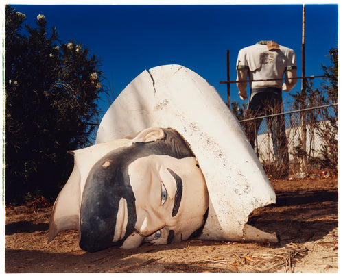 Photograph by Richard Heeps. In the foreground of the photograph is a broken off head of a giant fibreglass sculpture of a cowboy (a Muffler Man). In the background set in a deep blue sky is the muffler man's headless torso.