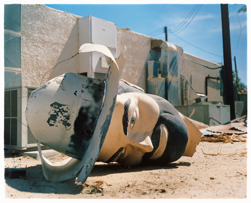 Photograph by Richard Heeps. The broken off head of a giant fibreglass sculpture of a cowboy (a Muffler Man) lies on the ground.