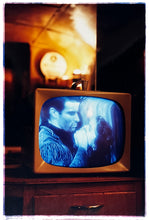 Load image into Gallery viewer, Photograph by Richard Heeps. A retro small brown television sits on a wooden table displaying a black and white film which shows as blue and white. In the background of the television is a warm light.