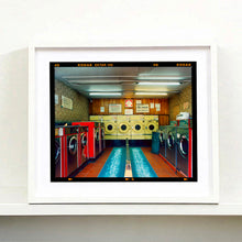 Load image into Gallery viewer, White framed photograph by Richard Heeps. A laundrette with washing machines on each wall and a double sided seat in the middle.
