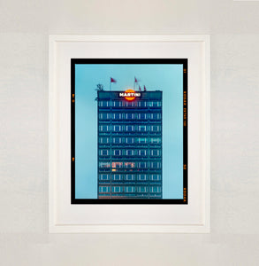 White framed photograph by Richard Heeps. High rise offices in a blue light with Martini logo on the top facade. 