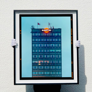 Black framed photograph held by photographer Richard Heeps. A photograph of high rise offices in a blue light with Martini logo on the top facade. 