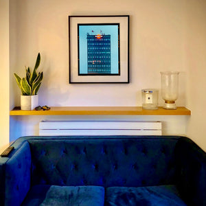 Black framed photograph in situ by photographer Richard Heeps. A photograph of high rise offices in a blue light with Martini logo on the top facade. 