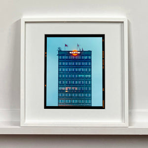 White framed photograph by Richard Heeps. High rise offices in a blue light with Martini logo on the top facade. 