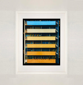 White framed photograph by Richard Heeps. Photograph of an apartment building with coloured balconies, blue at the top balcony and then fading from light yellow to a sunburnt yellow at the bottom.