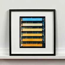 Load image into Gallery viewer, Black framed photograph by Richard Heeps. Photograph of an apartment building with coloured balconies, blue at the top balcony and then fading from light yellow to a sunburnt yellow at the bottom.