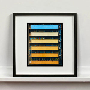 Black framed photograph by Richard Heeps. Photograph of an apartment building with coloured balconies, blue at the top balcony and then fading from light yellow to a sunburnt yellow at the bottom.