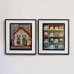 Two black framed photographs by Richard Heeps. On the left hand side is the photograph of a traditional Italian Tobacconist shop, here in a Swiss Cottage style building set against a vast urban apartment block. The right hand side photograph is of the balconies of a colourfully painted block of apartments.