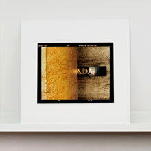 Load image into Gallery viewer, Mounted photograph by Richard Heeps. Brown flecked marble walls in different tones. In the middle is half a brown plaque with golden letters showing half an A, followed by a D and an A.