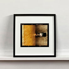 Load image into Gallery viewer, Black framed photograph by Richard Heeps. Brown flecked marble walls in different tones. In the middle is half a brown plaque with golden letters showing half an A, followed by a D and an A.