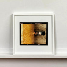 Load image into Gallery viewer, White framed photograph by Richard Heeps. Brown flecked marble walls in different tones. In the middle is half a brown plaque with golden letters showing half an A, followed by a D and an A.