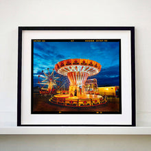 Load image into Gallery viewer, Black framed photograph by Richard Heeps. A fairground ride, the chairoplanes, sits lit in golden and red colours against a dark blue sky.