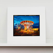 Load image into Gallery viewer, Mounted photograph by Richard Heeps. A fairground ride, the chairoplanes, sits lit in golden and red colours against a dark blue sky.