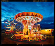 Load image into Gallery viewer, Photograph by Richard Heeps. A fairground ride, the chairoplanes, sits lit in golden and red colours against a dark blue sky.