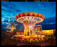 Photograph by Richard Heeps. A fairground ride, the chairoplanes, sits lit in golden and red colours against a dark blue sky.