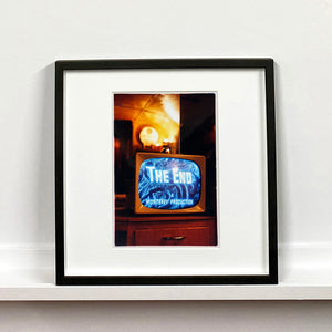 Black framed photograph by Richard Heeps. A retro small brown television sits on a wooden table displaying The End on a black and white film which shows as blue and white. In the background of the television is a warm light.