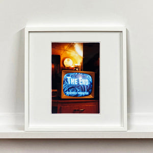 White framed photograph by Richard Heeps. A retro small brown television sits on a wooden table displaying The End on a black and white film which shows as blue and white. In the background of the television is a warm light.