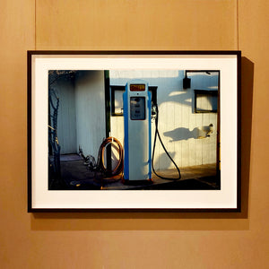 Black framed photograph by Richard Heeps. A vintage petrol pump with a white front and blue sides, sitting outside a white slatted building.