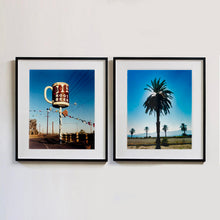 Load image into Gallery viewer, Two black framed photographs by Richard Heeps. A giant model of a mug with Bob&#39;s Root Beer written on it sits on top of a giant pole. There is bunting hanging from the pole. It sits alongside a power line on a remote looking American country road. The photograph on the right hand side features a palm tree with palm trees in the background, set in front of a blue sky and the sun filtering from behind the fronds of the tree.
