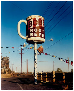 Photograph by Richard Heeps. A giant model of a mug with Bob's Root Beer written on it sits on top of a giant pole. There is bunting hanging from the pole. It sits alongside a power line on a remote looking American country road.