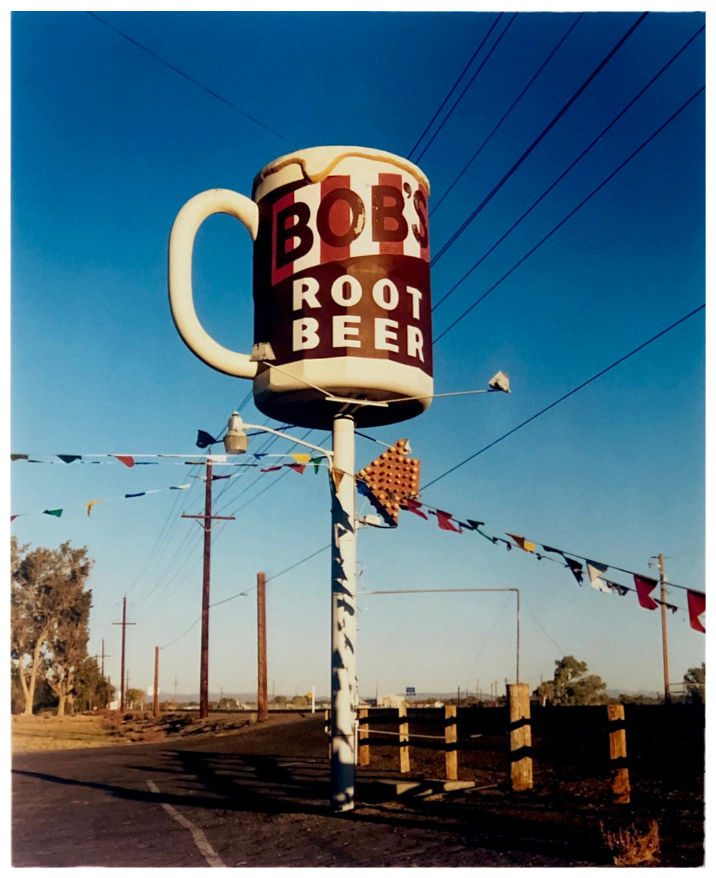 Photograph by Richard Heeps. A giant model of a mug with Bob's Root Beer written on it sits on top of a giant pole. There is bunting hanging from the pole. It sits alongside a power line on a remote looking American country road.