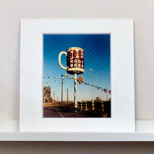 Load image into Gallery viewer, Mounted photograph by Richard Heeps. A giant model of a mug with Bob&#39;s Root Beer written on it sits on top of a giant pole. There is bunting hanging from the pole. It sits alongside a power line on a remote looking American country road.