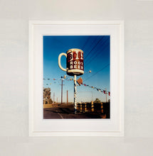Load image into Gallery viewer, White framed photograph by Richard Heeps. A giant model of a mug with Bob&#39;s Root Beer written on it sits on top of a giant pole. There is bunting hanging from the pole. It sits alongside a power line on a remote looking American country road.