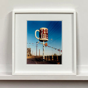 White framed photograph by Richard Heeps. A giant model of a mug with Bob's Root Beer written on it sits on top of a giant pole. There is bunting hanging from the pole. It sits alongside a power line on a remote looking American country road.