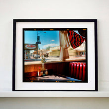 Load image into Gallery viewer, Black framed photograph by Richard Heeps. Inside an American diner the light is shining on the table and the seat is the classic vibrant red faux-leather of a retro diner. The photograph is looking out of the window into the outside street.