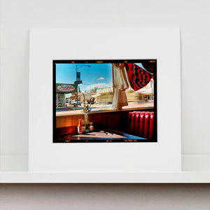 Mounted photograph by Richard Heeps. Inside an American diner the light is shining on the table and the seat is the classic vibrant red faux-leather of a retro diner. The photograph is looking out of the window into the outside street.