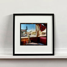Load image into Gallery viewer, Black framed photograph by Richard Heeps. Inside an American diner the light is shining on the table and the seat is the classic vibrant red faux-leather of a retro diner. The photograph is looking out of the window into the outside street.