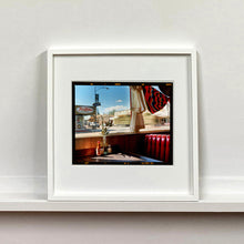 Load image into Gallery viewer, White framed photograph by Richard Heeps. Inside an American diner the light is shining on the table and the seat is the classic vibrant red faux-leather of a retro diner. The photograph is looking out of the window into the outside street.