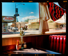 Load image into Gallery viewer, Photograph by Richard Heeps. Inside an American diner the light is shining on the table and the seat is the classic vibrant red faux-leather of a retro diner. The photograph is looking out of the window into the outside street.
