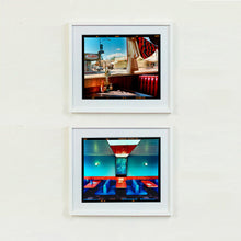 Load image into Gallery viewer, Two white framed photographs by Richard Heeps. The top one shows the Inside an American diner the light is shining on the table and the seat is the classic vibrant red faux-leather of a retro diner. The photograph is looking out of the window into the outside street. The bottom photograph is the inside of a Vintage Wimpy in Norfolk, bright blue retro seating and red tables, set against a blue wall.