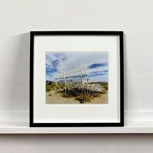 Load image into Gallery viewer, Black framed photograph by Richard Heeps. The back view of a basic white painted grandstand sits in the middle of this photograph. Grass grows up from its base and it sits alone on a sandy ground.