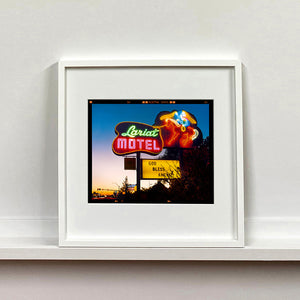 White framed photograph by Richard Heeps. The photograph is taken in the evening, a neon sign belonging to the Lariat Motel, with a cowboy on horseback with a lasso on the right of the words, half way down the sign post is the yellow sign God Bless America.