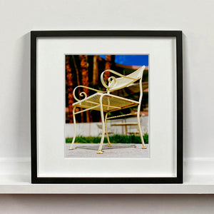 Black framed photograph by Richard Heeps. A cream chair sits on hard standing, in the back and slightly out of focus is lush green grass and warm red tree trunks.