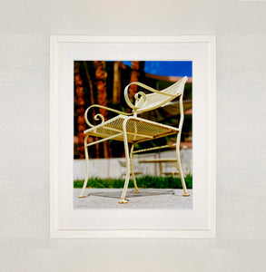 White framed photograph by Richard Heeps. A cream chair sits on hard standing, in the back and slightly out of focus is lush green grass and warm red tree trunks.