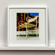Load image into Gallery viewer, White framed photograph by Richard Heeps. A cream chair sits on hard standing, in the back and slightly out of focus is lush green grass and warm red tree trunks.