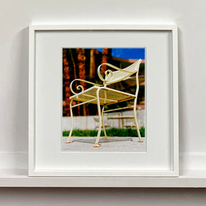White framed photograph by Richard Heeps. A cream chair sits on hard standing, in the back and slightly out of focus is lush green grass and warm red tree trunks.