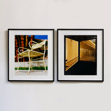 Load image into Gallery viewer, Two black framed photographs by Richard Heeps. The photograph on the left is of a cream chair sitting on hard standing, behind the chair and slightly out of focus is lush green grass and warm red tree trunks. The photograph on the right hand side depicts the evening sun casting a shadow as it glows through a window in a door.