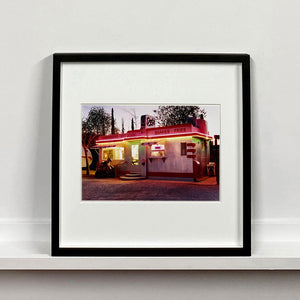Black framed photograph by Richard Heeps. This photograph depicts a one storey small building "Dot's Diner" brightly lit with a pink roof, with Hamburgers, Hot Dogs, Shakes, Fries written along the top width of the building.